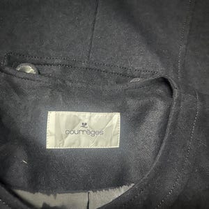 May include: Close-up of a dark navy blue jacket with a suede-like texture. The jacket features a small pocket with a button and a label that reads "courr&egrave;ges". The stitching is visible, and the overall design is minimalist.