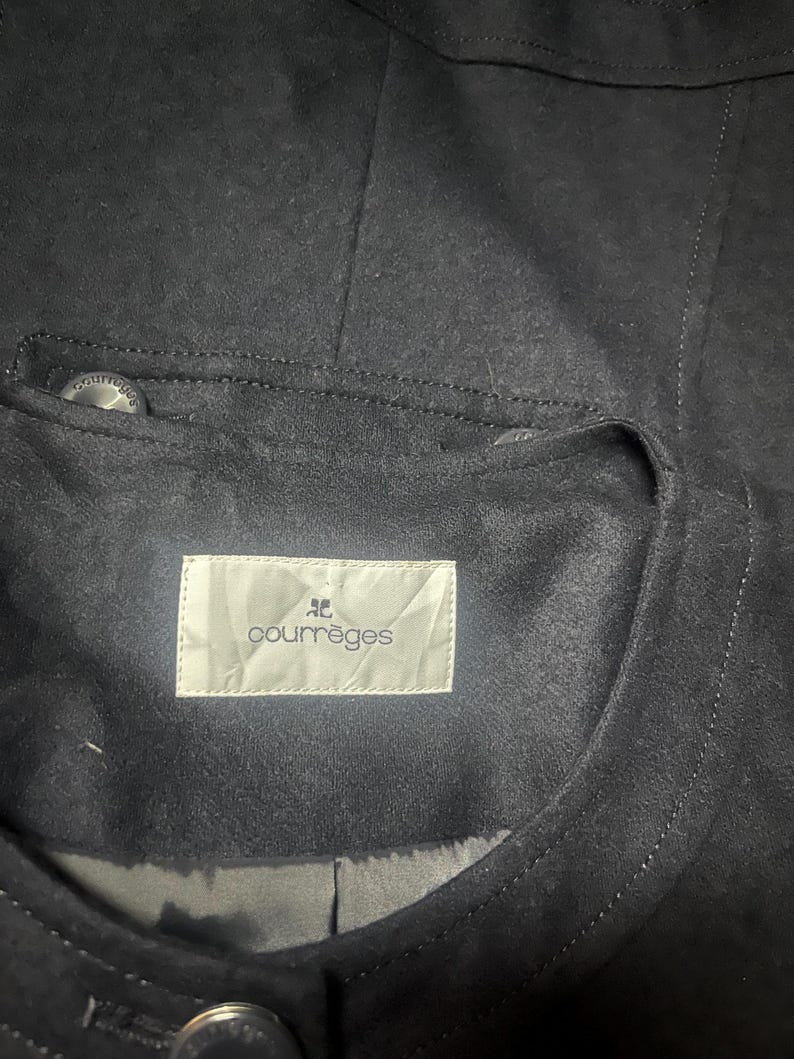 May include: Close-up of a dark navy blue jacket with a suede-like texture. The jacket features a small pocket with a button and a label that reads "courr&egrave;ges". The stitching is visible, and the overall design is minimalist.