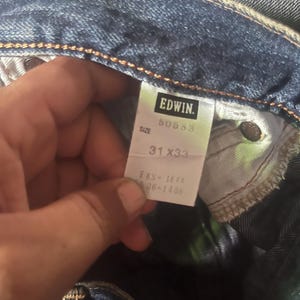 May include: Close-up of blue denim jeans with a tag that reads "EDWIN." The tag also displays the size as 31x33. The jeans are folded, showing the inside pocket and stitching details.