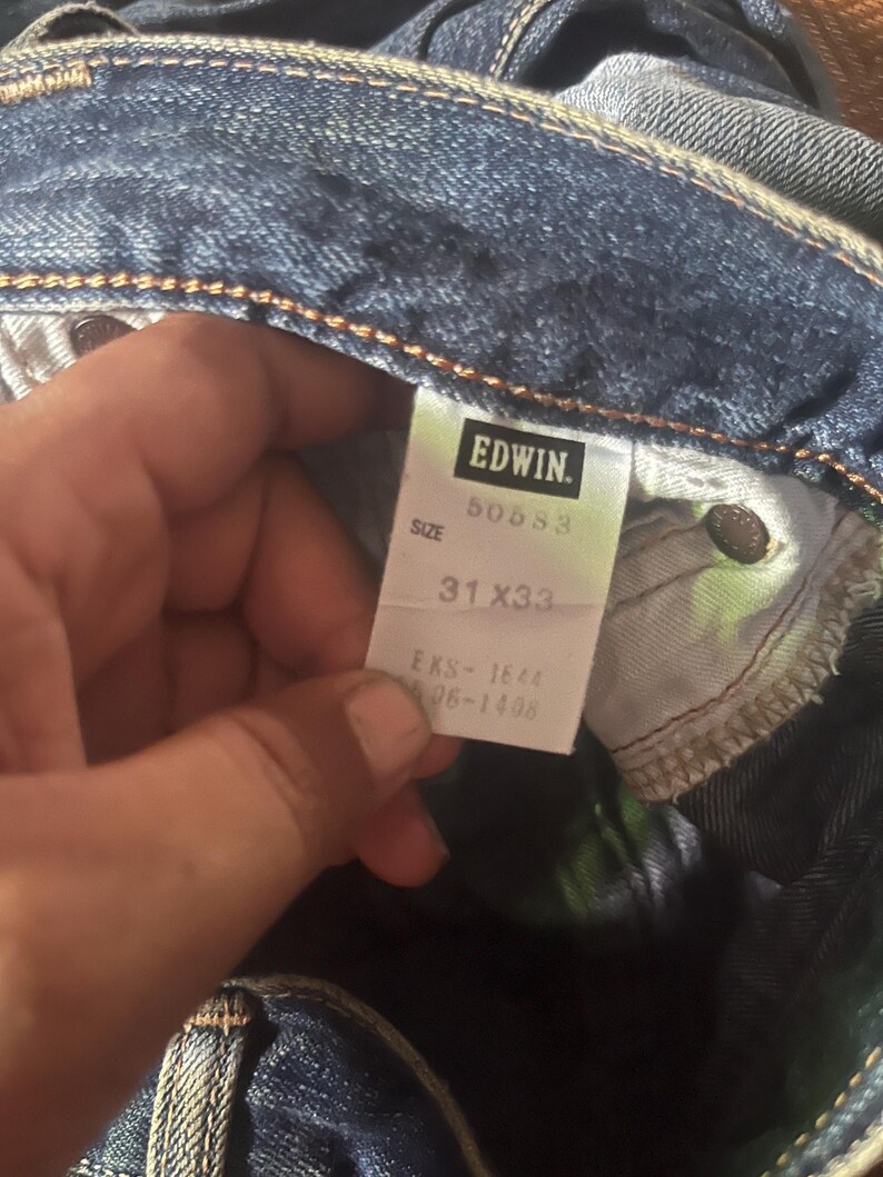 May include: Close-up of blue denim jeans with a tag that reads "EDWIN." The tag also displays the size as 31x33. The jeans are folded, showing the inside pocket and stitching details.