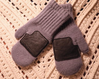 Könnte beinhalten: Ein Paar graue Strickhandschuhe mit schwarzen Lederflicken auf den Handflächen. Die Handschuhe liegen auf einer weißen Häkelunterlage.