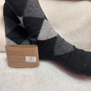 May include: A pair of black, gray, and charcoal argyle socks with thin red accent lines. A brown paper label is attached, featuring text about alpaca fiber, care instructions, and a white sticker with the text 7220 Black M 41% Baby Alpaca.