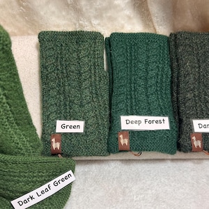 May include: Four pairs of fingerless gloves in various shades of green, including Dark Leaf Green, Green, Deep Forest, and Dark Green. Each pair features a cable knit pattern and a small brown tag with a white llama icon. Labels identify each color.