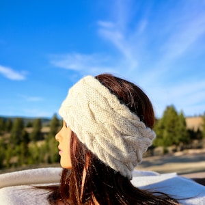 May include: A white knitted headband with a cable knit pattern. The headband is worn by a person with long brown hair. The background is a blue sky with clouds.