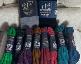 Puede incluir: Una colección de calcetines coloridos, incluyendo verde, negro, gris, morado, marrón, rojo, verde azulado y azul. Los calcetines están empaquetados y etiquetados con el texto "New England Woolens" y "All Day Alpaca Crew Socks".