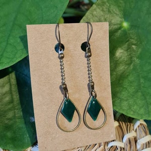 May include: A pair of dangle earrings with a green diamond-shaped charm and a gold teardrop-shaped frame. The earrings are suspended from a chain with a gold hook.