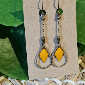 May include: A pair of earrings with a teardrop-shaped frame and a bright yellow diamond-shaped insert. The earrings have a chain and hook design, with a bronze finish. The earrings are displayed on a brown card.