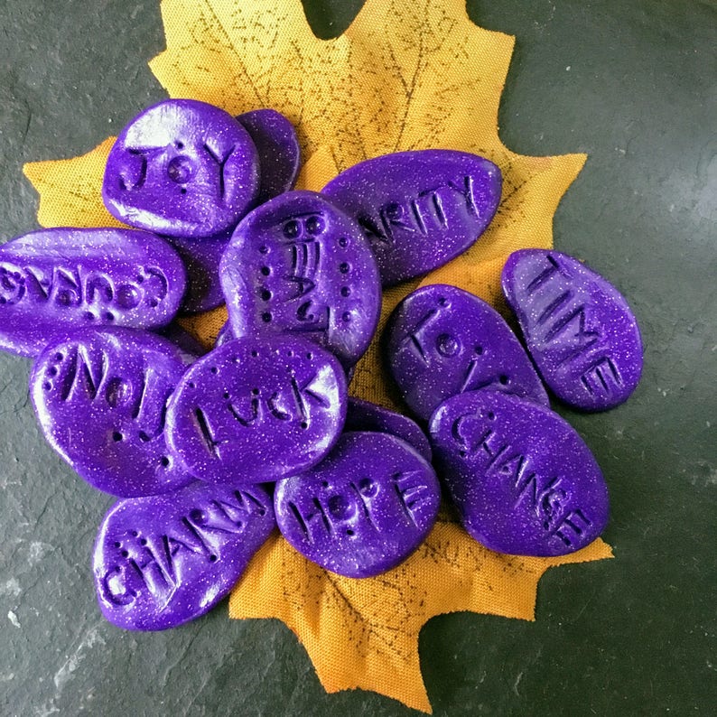 May include: Purple painted stones with words such as "Joy", "Time", "Hope", "Luck", and "Change" written on them. The stones are arranged on a yellow leaf.
