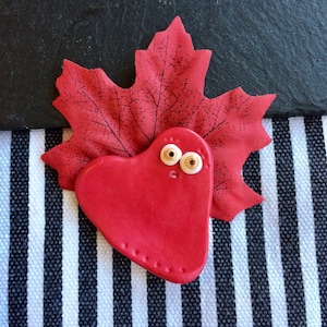 May include: A red heart-shaped ceramic ornament with googly eyes, sitting on a red maple leaf. The leaf is on a black and white striped background.