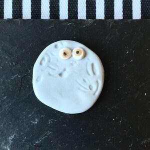 May include: A circular, light blue clay object with a smooth, matte finish. It features two small, round, beige-coloured eyes with dark centres positioned near the top. The surface has subtle, shallow indentations and markings across its face.