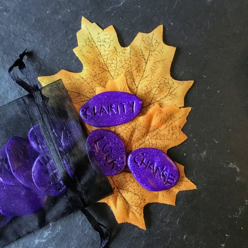 May include: Purple and black stones with the words "Clarity", "Luck", and "Change" written on them. The stones are in a black mesh bag and on top of a yellow leaf.