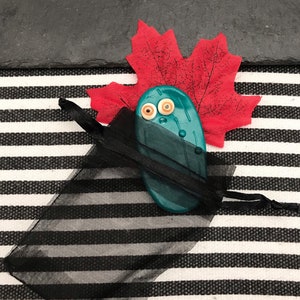 May include: A teal, oval-shaped clay figure with two small, round, white and orange button eyes, partially tucked inside a sheer black drawstring organza pouch, positioned in front of a single, vibrant red artificial maple leaf.