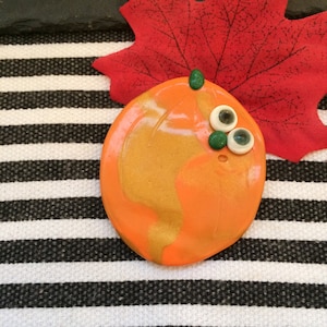 May include: A smooth, oval-shaped orange and gold pumpkin-like object featuring a small green stem at the top. It has two white googly eyes and a small green circular accent attached to the front surface, creating a simple, rounded character design.