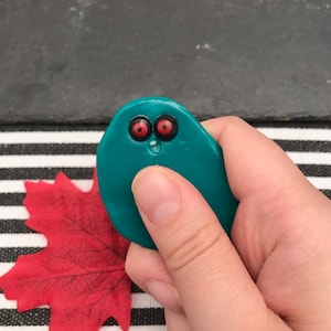 May include: A smooth, teal-coloured, rounded object with two prominent red eyes featuring black pupils and a small, white, open-mouthed expression. The object has a glossy finish and is held in place, showcasing its compact, pebble-like shape and vibrant colour.
