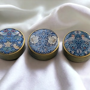 Può includere: Tre scatole rotonde per gioielli con bordi dorati e motivi floreali blu e bianchi. I disegni presentano uccelli e fiori stilizzati.