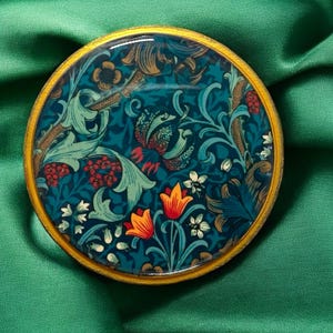 May include: A round coaster with a gold rim, featuring a teal floral design with orange and white flowers, green leaves, and red berries. The coaster is set against a green fabric background.