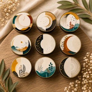 May include: A set of nine circular metal tins, each featuring a unique artistic design on the lid. The illustrations include celestial motifs like moons and suns, alongside botanical elements, mountain landscapes, and abstract patterns in earthy tones.