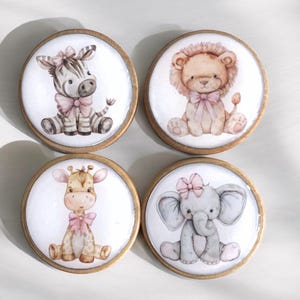 Peut inclure: Quatre boutons en bois avec un fond blanc et une image peinte d'un zèbre, d'un lion, d'une girafe et d'un éléphant. Chaque animal porte un nœud rose.