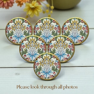 May include: Six round decorative cabinet knobs with a floral design. Each knob features a white background with a symmetrical pattern of yellow, red, blue, and green floral elements, framed by a gold-colored edge. The text "Please look through all photos" is at the bottom.
