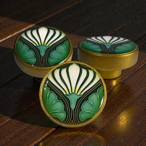 Könnte beinhalten: Drei dekorative Schrankknöpfe mit floralem Design. Die Knöpfe haben ein weißes und grünes Blumenmuster mit goldfarbenem Rand. Die Knöpfe sind auf einer dunklen Holzoberfläche platziert.
