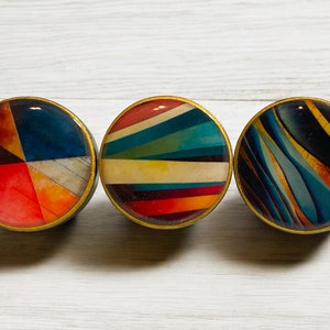 May include: Three round cabinet knobs with colorful abstract designs. Each knob has a gold-colored rim and a unique pattern: one with red, orange, and blue segments; another with striped bands; and a third with curved lines.
