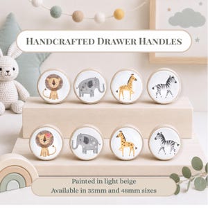 Op de afbeelding: Acht ronde houten ladeknoppen met lichtbeige randen en witte centra met illustraties van een leeuw, olifant, giraf en zebra. De tekst luidt Handcrafted Drawer Handles, Painted in light beige, Available in 35mm and 48mm sizes.