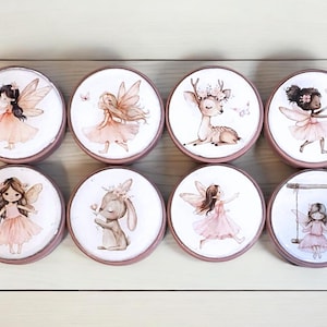 Może przedstawiać: Osiem okrągłych drewnianych pudełek z różowymi pokrywkami. Każde pudełko zawiera inną ilustrację wróżki, jelenia, królika i huśtawki. Ilustracje są w stylu akwarelowym w kolorze różowym, białym i brązowym.