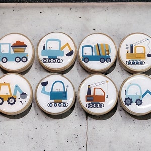 Pode incluir: Conjunto de 8 ímãs redondos de madeira com diferentes veículos de construção em azul, amarelo, laranja e marrom. Cada ímã tem um fundo branco com contornos pretos.