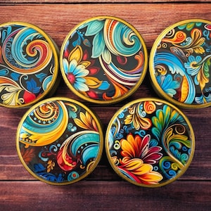 Op de afbeelding: Vijf ronde, decoratieve onderzetters met een gouden rand. Elke onderzetter heeft een ander kleurrijk bloemmotief op een zwarte achtergrond.