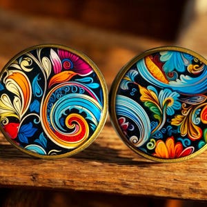 May include: Two round, gold-rimmed, decorative buttons with a colorful, swirling floral pattern on a dark background. The pattern includes shades of blue, red, yellow, green, and orange.