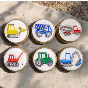 May include: Six round wooden knobs with different construction vehicle designs. The knobs feature a yellow bulldozer, a blue dumper truck, a red cement mixer, a red crane, a green tractor, and a blue excavator.