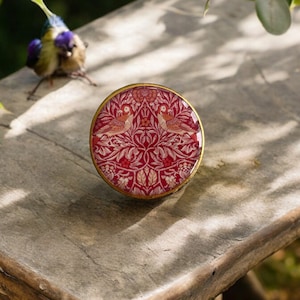 May include: A round, decorative trinket box with a red and white floral and bird design. The box has a gold-colored rim and a glossy finish. The box is sitting on a wooden surface.