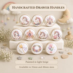 Puede incluir: Un juego de once tiradores redondos para cajones con ilustraciones de unicornios, un castillo, un carruaje, una varita de estrella, una corona floral y globos. El texto dice HANDCRAFTED DRAWER HANDLES, Painted in light beige, 35mm y 48mm.