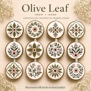 Pu&ograve; includere: Dodici pomelli per cassetti rotondi in ceramica etichettati da A a L, ognuno con motivi unici di foglie di olivo e fiori in tonalit&agrave; di verde e rosa su sfondo bianco, disposti in una griglia con il testo Olive Leaf Handcrafted Decorative Drawer Knobs.