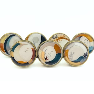 Peut inclure: Six boutons de porte ronds en or avec un fond blanc et des motifs abstraits dans des tons de bleu, marron et orange.