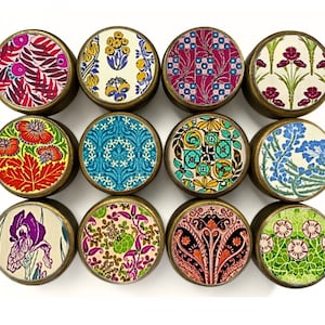 May include: A collection of twelve round decorative knobs with brass-colored metal rims. Each knob features a different colorful floral or patterned design on its top surface, including leaves, flowers, and geometric shapes. The knobs are arranged in a grid.