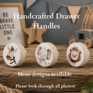May include: Three round drawer handles with a light beige base and a flat white top. Each handle features a different animal illustration: a zebra on the left, an elephant in the centre, and a lion on the right. The text reads Handcrafted Drawer Handles.
