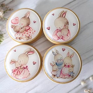 May include: Four round wooden knobs with gold trim, each featuring a different hand-painted illustration of a bunny family. The bunnies are depicted in various poses, including holding hands, hugging, and carrying a soccer ball. The knobs are perfect for adding a touch of whimsy to any room.