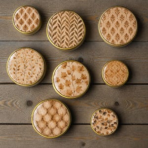 Op de afbeelding: Acht ronde metalen blikjes met goudkleurige randen, elk met een uniek bruin en beige patroon op het deksel. De patronen bevatten diverse botanische motieven zoals bladeren, esdoornbladeren en geometrische ontwerpen op een plat oppervlak.