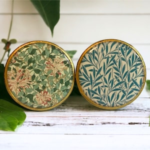 Pode incluir: Dois botões redondos com aro dourado e padrões florais. Um botão apresenta um desenho floral verde e branco com flores cor-de-rosa. O outro botão apresenta um desenho floral azul e branco.