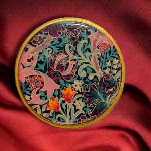 May include: A round, decorative compact with a gold rim. The compact features a floral design in shades of teal, pink, purple, and orange. The floral pattern includes lilies and other stylized flowers. The background is a deep red.