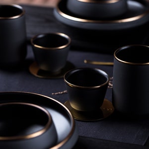 May include: A set of black ceramic cups and saucers with gold rims. The cups and saucers are arranged on a black surface.