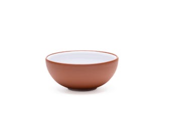 Puede incluir: Un cuenco pequeño y redondo con un exterior liso de color terracota mate y un interior blanco brillante que contrasta. El cuenco presenta una silueta sencilla y curva con una abertura ancha y una base plana, ofreciendo un diseño limpio.