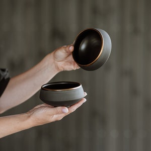 May include: Two black ceramic bowls with a gold rim. The bowls are nested together, with the top bowl slightly tilted.