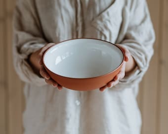 Puede incluir: Una persona sosteniendo un tazón de terracota con un interior blanco. El tazón es redondo y tiene un borde ligeramente acampanado.