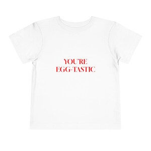 May include: A white t-shirt with the red text "YOU'RE EGG-TASTIC" printed on the front. The shirt is a short-sleeved, crew neck style, suitable for children.