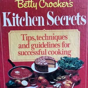 May include: A cookbook cover titled "Betty Crocker's Kitchen Secrets" in yellow and white text on a red background. The cover features illustrations of food items, including a steak in a pan, a cake, and vegetables.