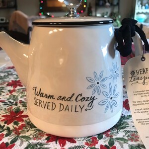 May include: White ceramic teapot with a black lid and a black ribbon handle. The teapot has a blue floral design and the text "Warm and Cozy SERVED DAILY" on the front.
