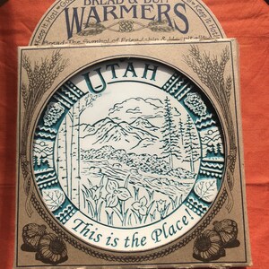 May include: A brown cardboard box with a white ceramic bread warmer inside. The warmer is round and features a blue and white design of a mountain scene with the word "UTAH" and the text "This is the Place!" The box has the text "Bread & Bun Warmers" on the top.