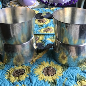 May include: Two sets of silver stacking cups with heart-shaped handles. The cups are stacked on top of each other and are sitting on a blue and yellow floral placemat.